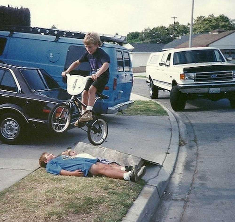 A child on a bicycle jumps over another child lying on the grass next to a curb, with parked cars and houses in the background.