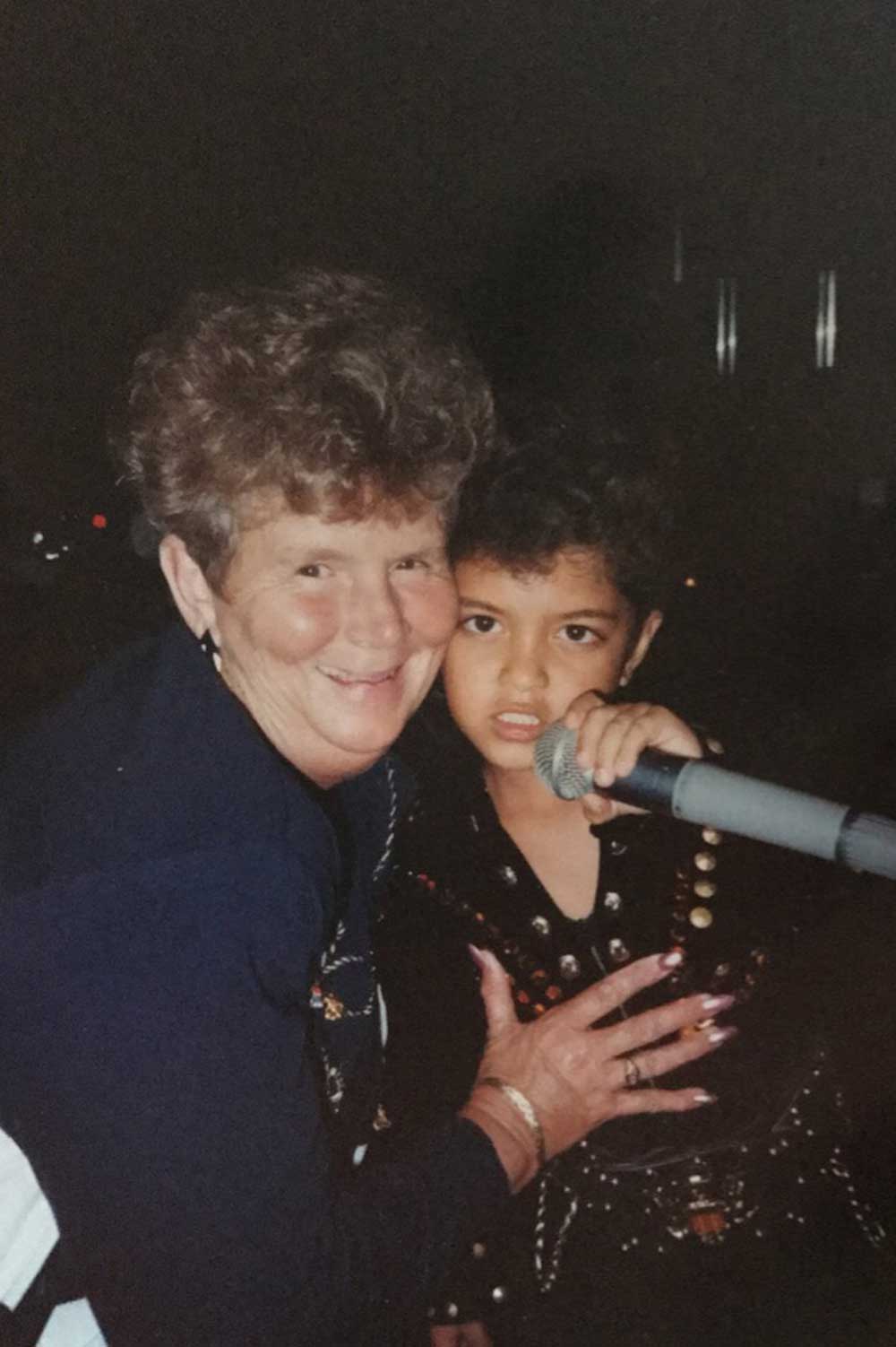 An older woman smiles and hugs a young boy dressed in a black, studded outfit holding a microphone. They are close together, with a dark background behind them.