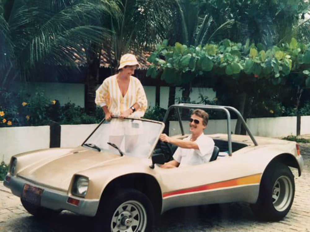 A man sits in a beige dune buggy with a red and yellow stripe, smiling up at a woman standing beside the car. They are outdoors, surrounded by greenery and palm trees, and both appear relaxed and happy.