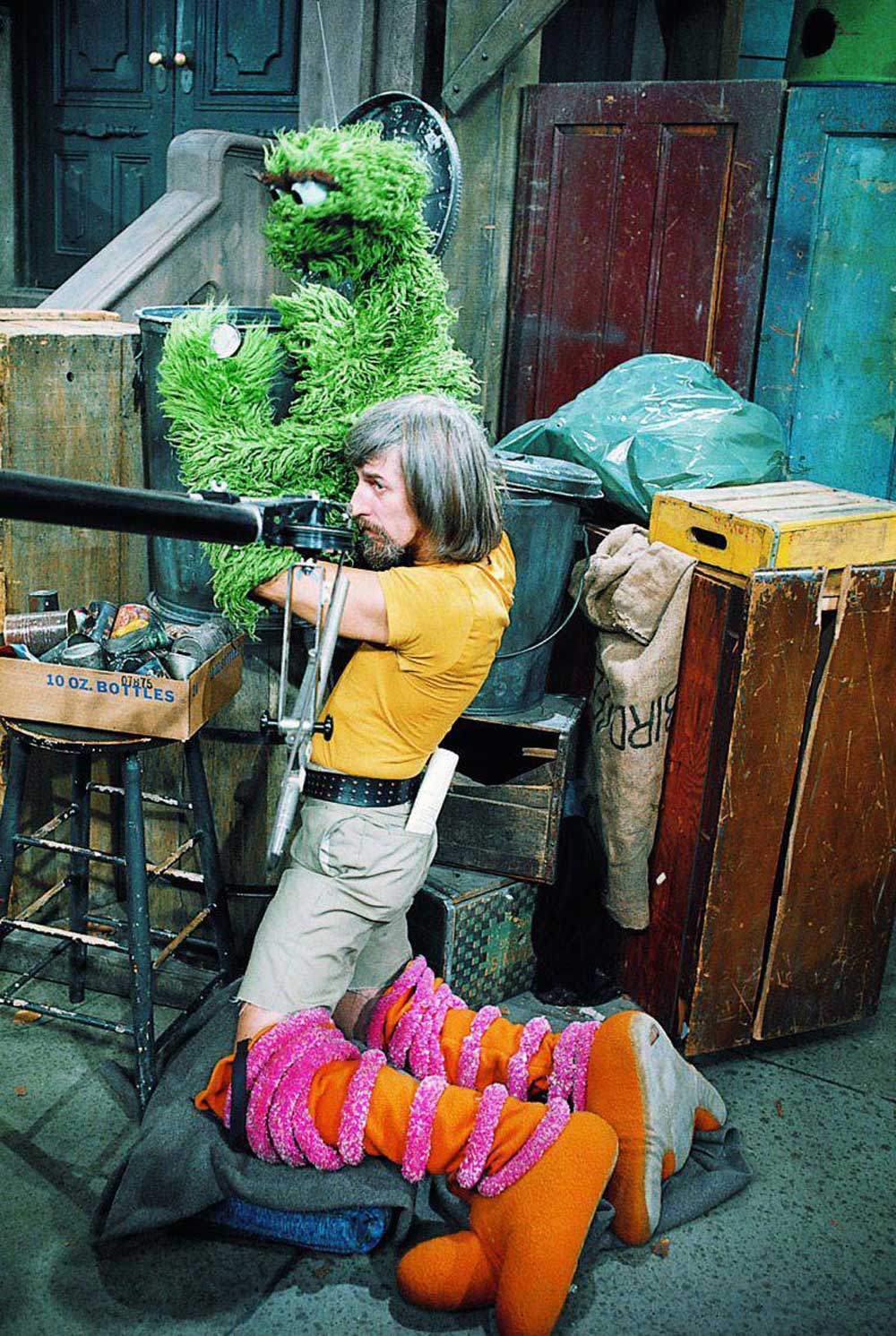 A puppeteer operates Oscar the Grouch from inside a trash can while sitting on the floor, surrounded by production equipment and props, with another puppet lying in the foreground.