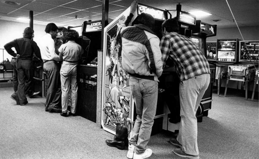 A group of people in casual 1980s clothing play arcade games in a dimly lit arcade. Some stand closely together, focused on the machines, while others wait nearby. The scene feels lively and nostalgic.