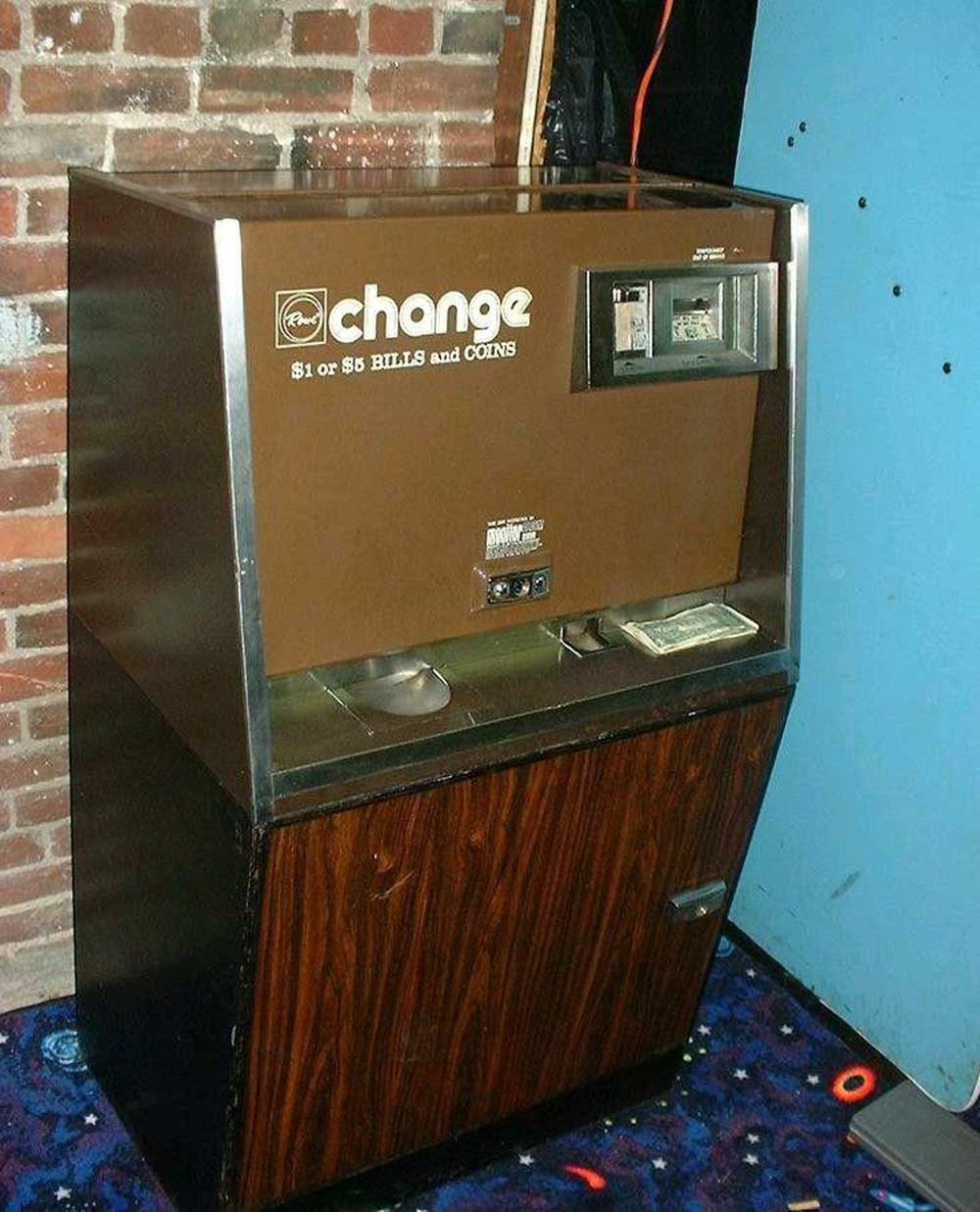 A vintage change machine against a brick wall, with a brown front panel labeled change for $1 or $5 bills and coins, and a wood-patterned lower section on a carpeted floor.