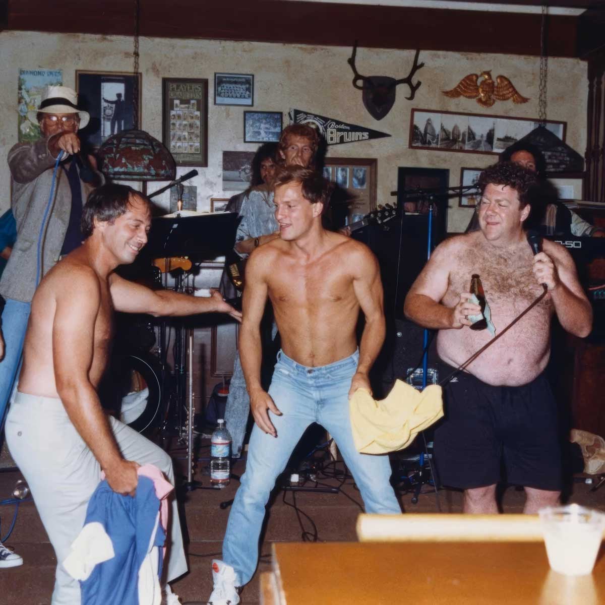 Three men, two of them shirtless and one holding a drink, dance and smile in a lively bar with a live band playing in the background. The scene is energetic and casual, with musical instruments and decorations visible.