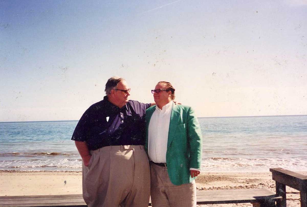 Two men stand side by side on a beach, smiling at each other. One wears a dark shirt and beige pants; the other wears a green blazer and sunglasses. The ocean and clear sky are visible in the background.