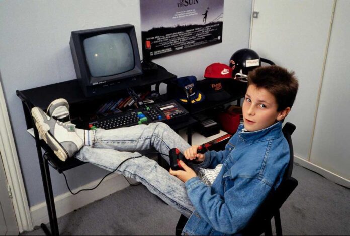 A boy in a denim jacket and jeans sits at a desk with one leg up, holding a joystick and looking at the camera. A vintage computer, caps, and other items are on the desk, with a poster on the wall behind.