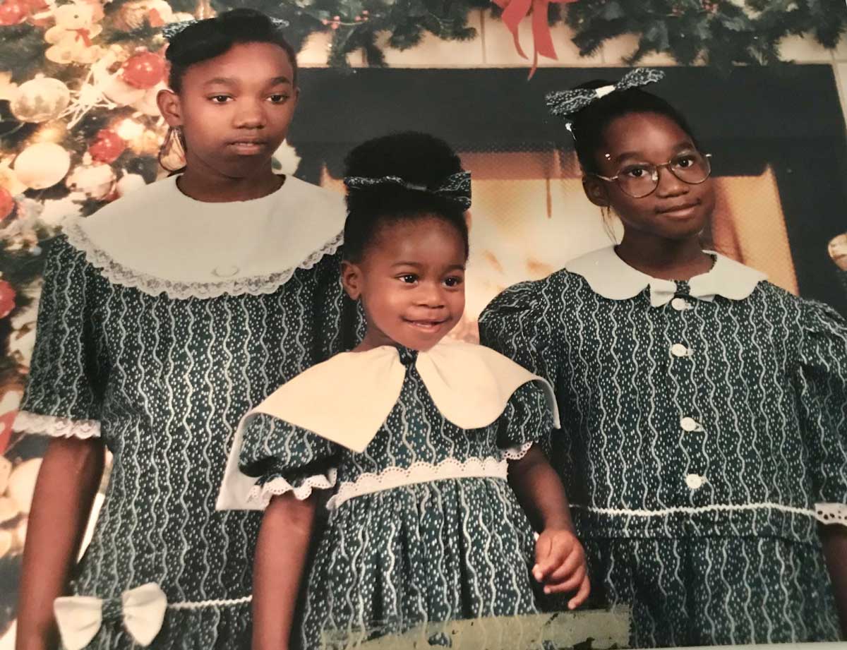 Three young girls in matching green and white dresses pose in front of a fireplace and decorated Christmas tree. The eldest and youngest smile, while the middle girl looks serious. All have white collars and hair bows.