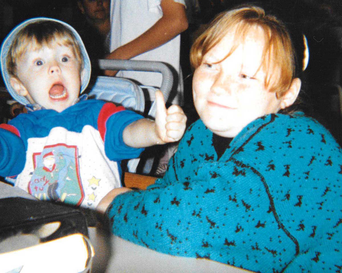 A young boy in a colorful hoodie makes a surprised face, giving a thumbs up, while a red-haired girl beside him smiles softly, both sitting indoors.