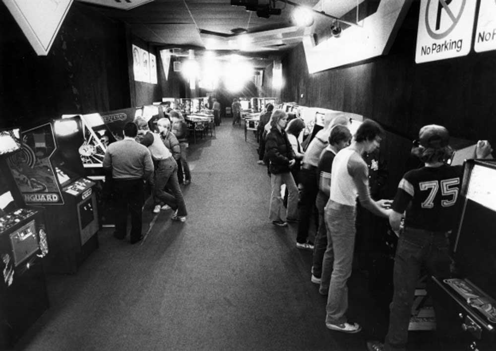 Black and white photo of a crowded 1980s arcade with people standing at rows of arcade machines on both sides of a long room. Several No Parking signs are visible on the dark walls.