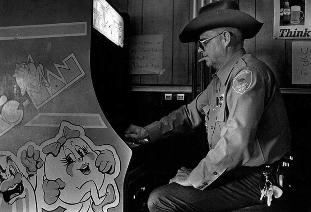 A man in a sheriff’s uniform and cowboy hat sits and plays a Ms. Pac-Man arcade game in a dimly lit room, with keys hanging from his belt and a cigarette in his mouth.