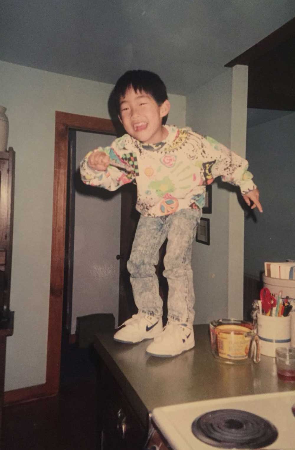 A young child in a colorful sweater and jeans stands laughing on a kitchen counter. There are household items and a stove visible in the foreground, with a doorway and pale walls in the background.
