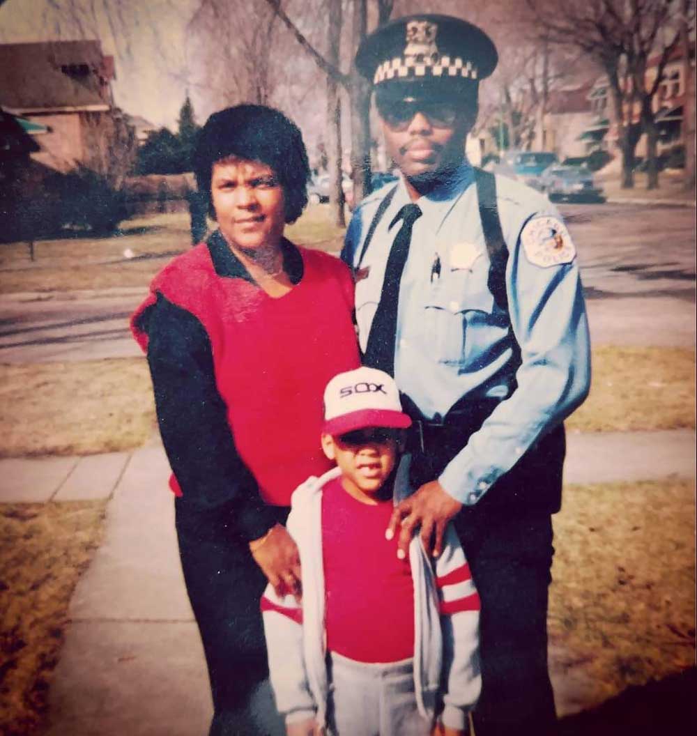 A woman, a young boy in a White Sox cap, and a man in a police uniform stand together on a sidewalk in a suburban neighborhood on a sunny day.
