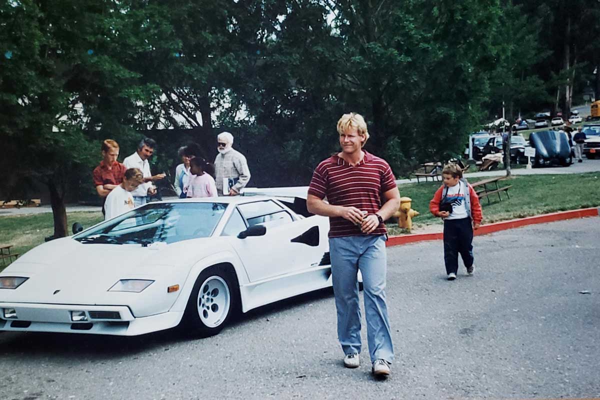 A man in a red striped shirt and blue pants walks in front of a white Lamborghini Countach parked on a street, with people and trees in the background at a park or outdoor gathering.
