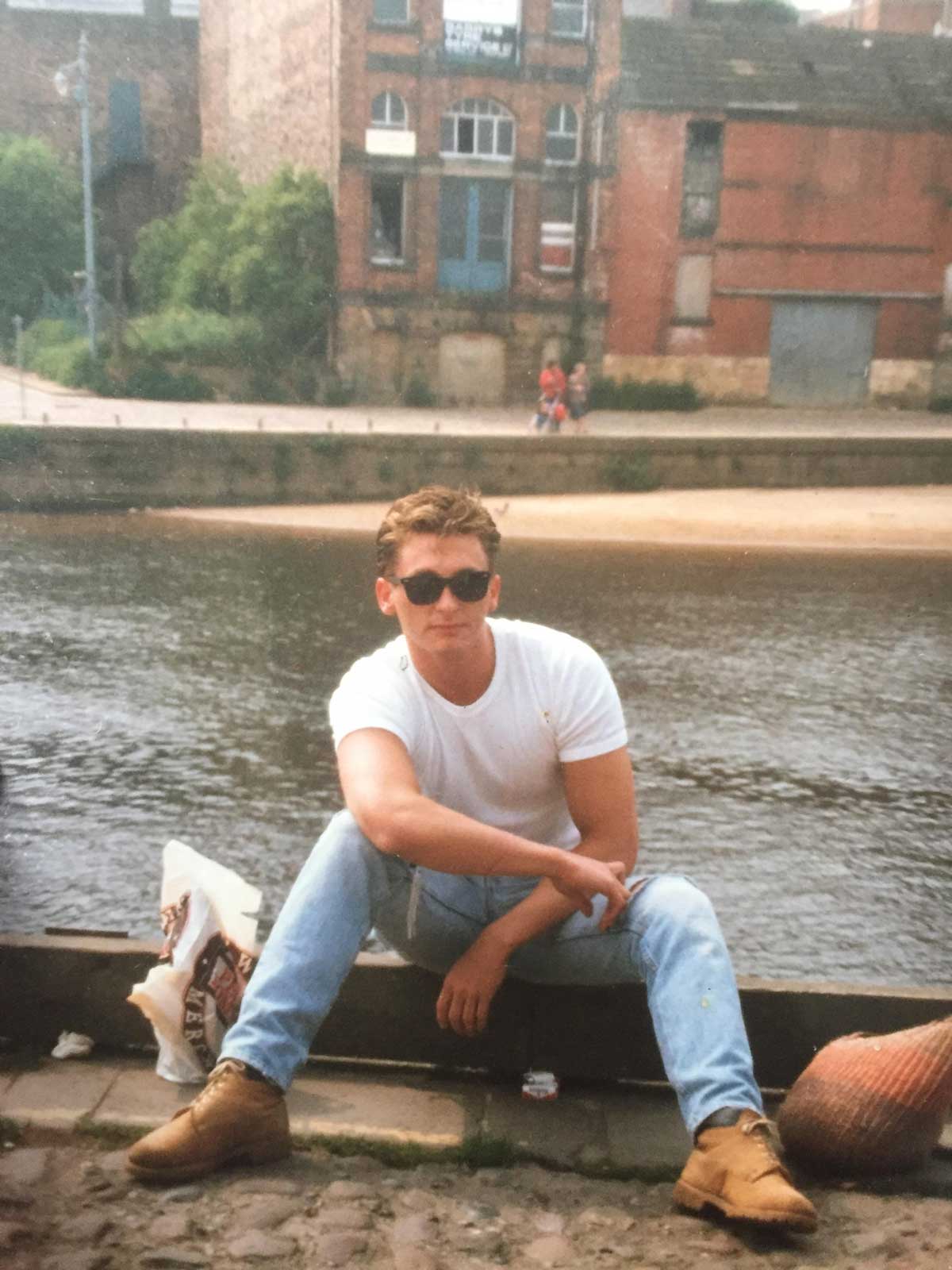 A young man wearing sunglasses, a white t-shirt, jeans, and brown boots sits casually by a riverbank with old brick buildings in the background. He has one arm resting on his knee and a plastic bag beside him.