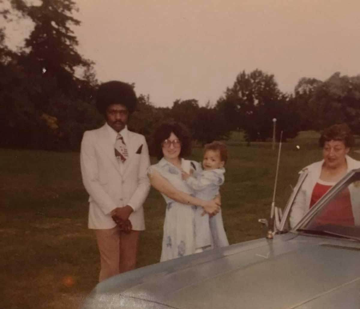 Four adults and a baby stand outdoors near a blue car. One woman holds the baby, while two others and a man in a white blazer stand nearby. Trees and grass fill the background.