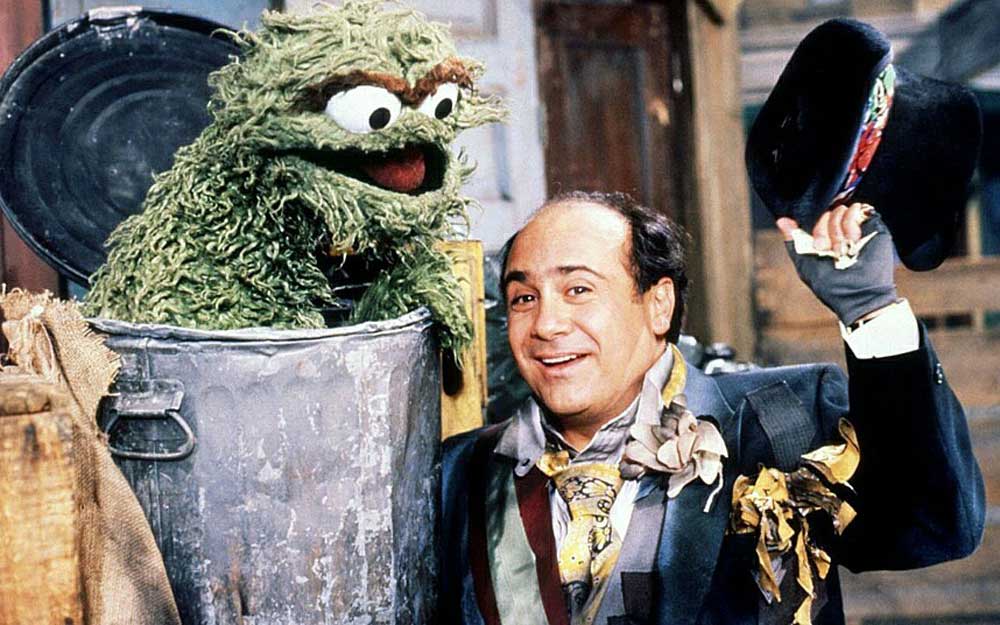 A man in a shabby suit smiles and tips his hat while standing next to Oscar the Grouch, a green puppet character in a trash can, on a street set resembling Sesame Street.