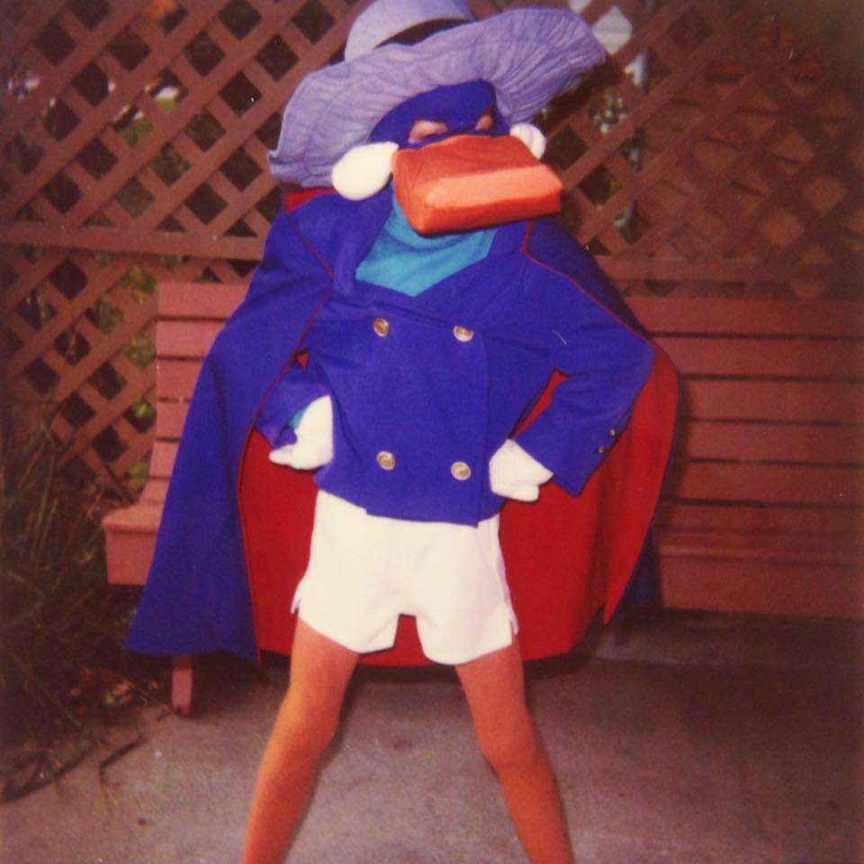 A person wearing a homemade Darkwing Duck costume, featuring a large purple hat, blue mask, beak, blue coat, white shorts, and white gloves, stands confidently on a sidewalk in front of a bench and wooden lattice.