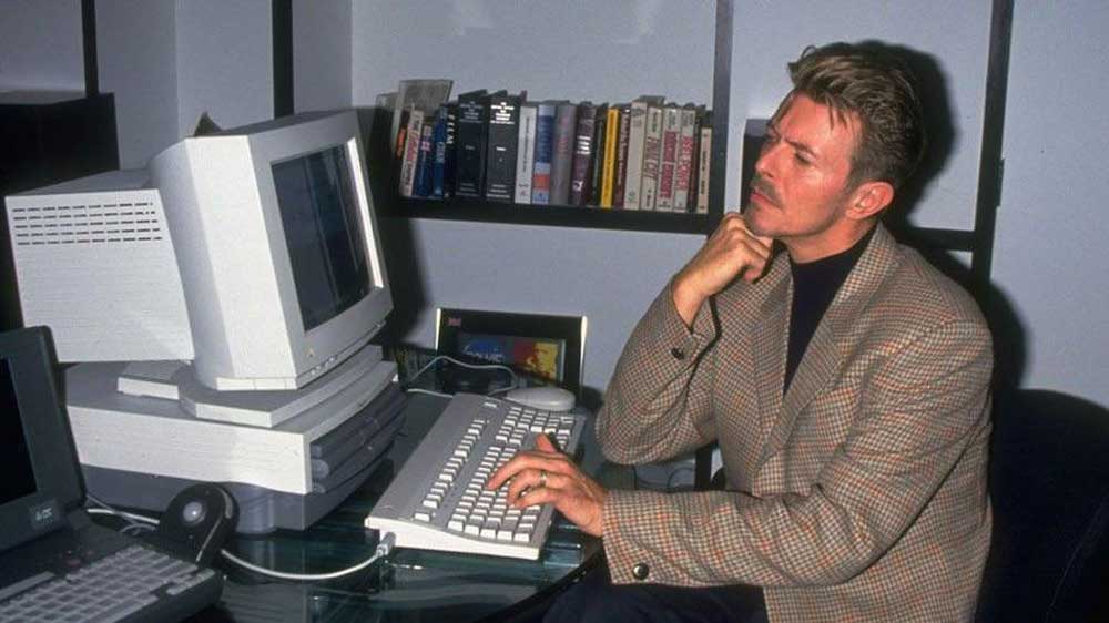 David Bowie in a plaid blazer sits at a desk, thoughtfully looking at a large, vintage computer monitor with one hand on the keyboard. Books and framed photos are visible on shelves behind him.