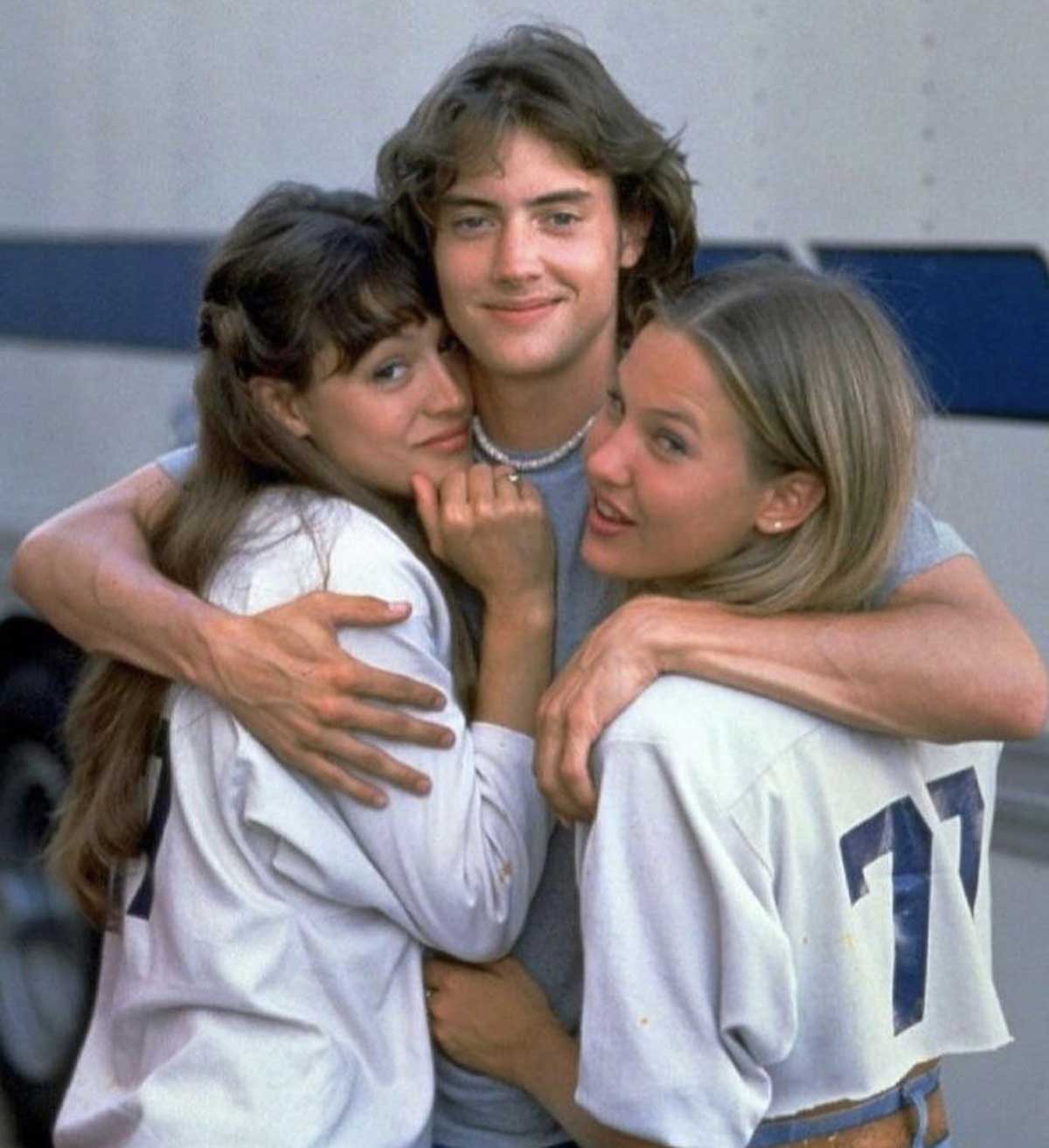 Three smiling teenagers stand close together. The boy in the middle hugs the two girls on either side. They appear happy and casual, with the girls wearing sports jerseys and the boy in a gray shirt.