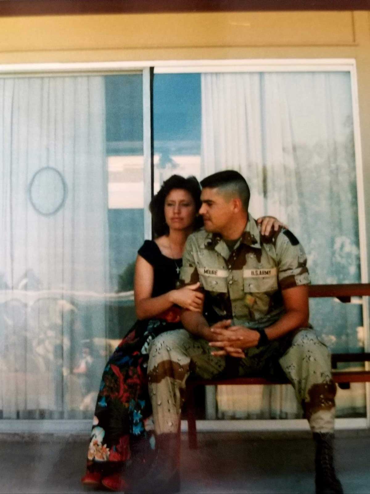 A woman sits beside a man in military uniform on a bench in front of a sliding glass door. She has her arm around his shoulders, and they both look ahead with serious expressions.