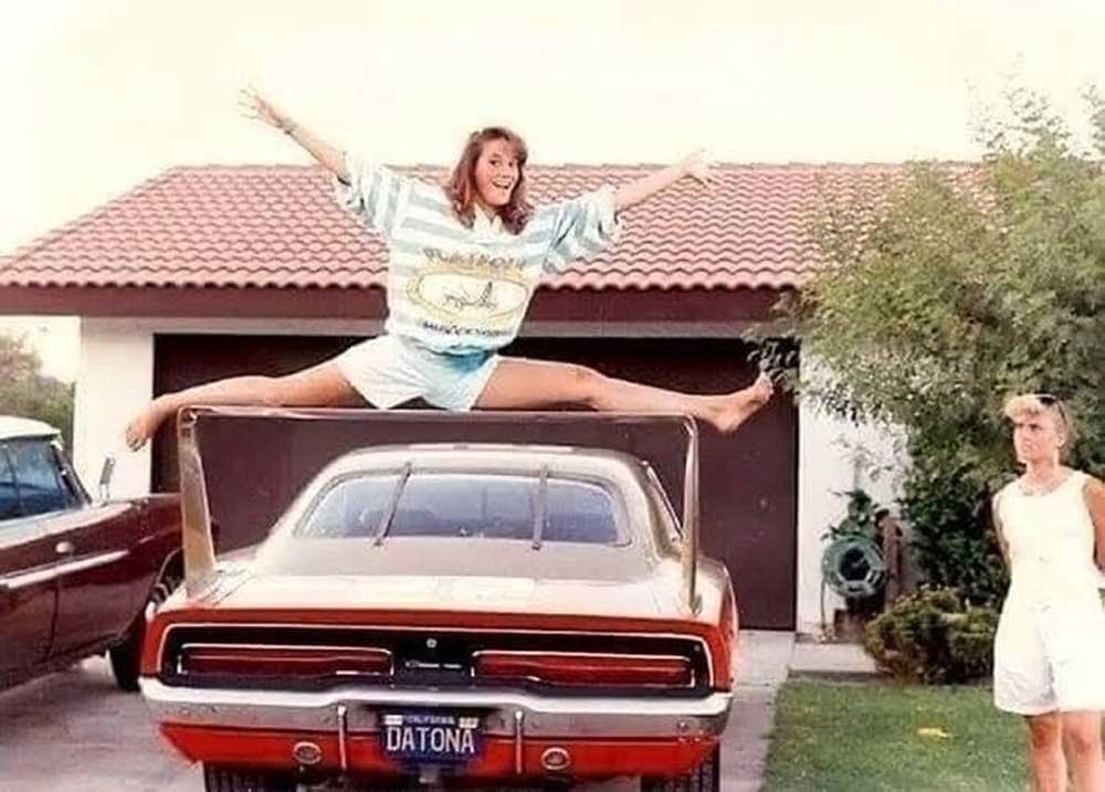 A woman does the splits on the rear spoiler of a red sports car parked in front of a house, while another woman stands nearby, watching. The car has a DAYTONA license plate.