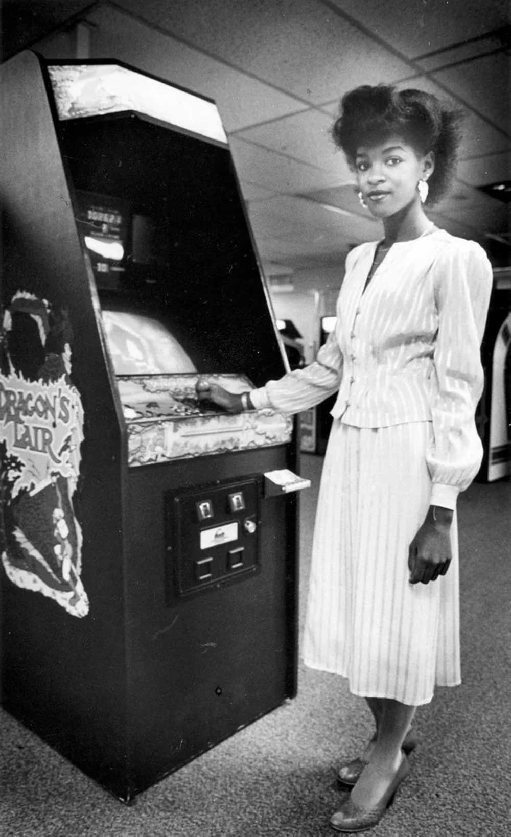A woman in a striped blouse and pleated skirt stands beside a Dragon’s Lair arcade machine in a vintage arcade, with one hand resting on the game controls.