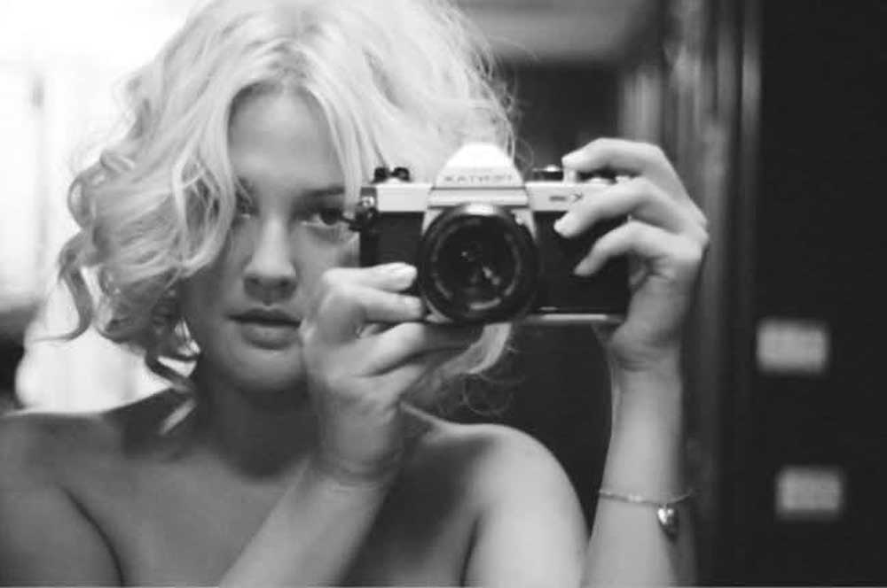 A person with short, curly blonde hair holds a vintage camera up to their face, looking into a mirror. The image is in black and white, and the person appears to be taking a self-portrait.