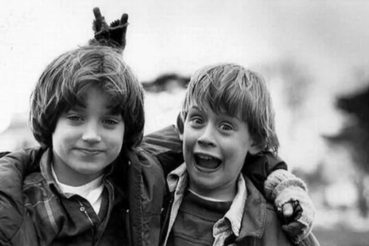 Two young boys pose together outdoors; one gives a playful smirk while the other looks surprised. The boy on the left has his arm around the other and holds up two fingers behind his friend’s head, making bunny ears.