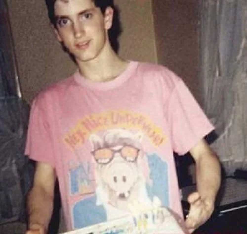 A young man wearing a pink T-shirt with a cartoon character design stands indoors, holding a pizza box and smiling at the camera.