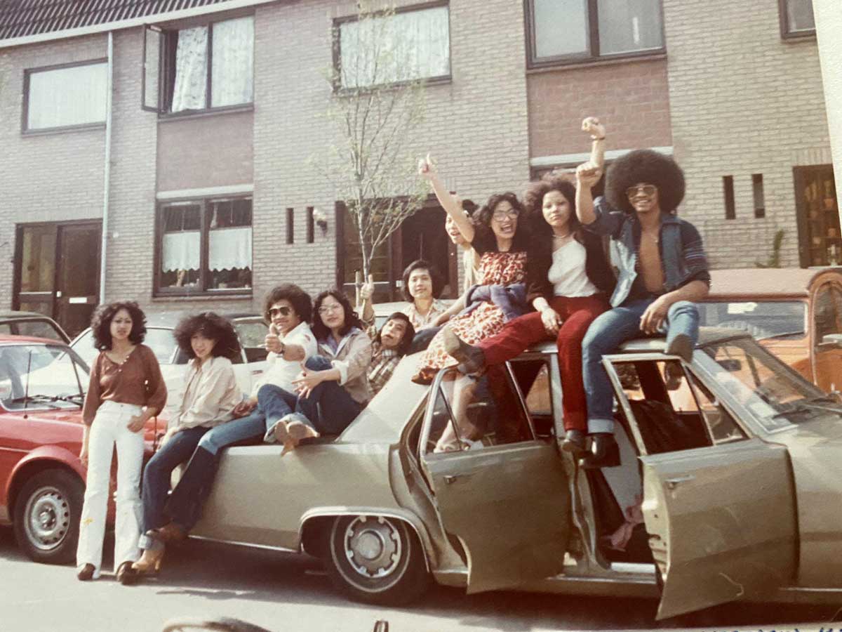 A group of young people with big hairstyles sit and pose cheerfully on and around a tan car parked on a residential street, with houses and other cars in the background.