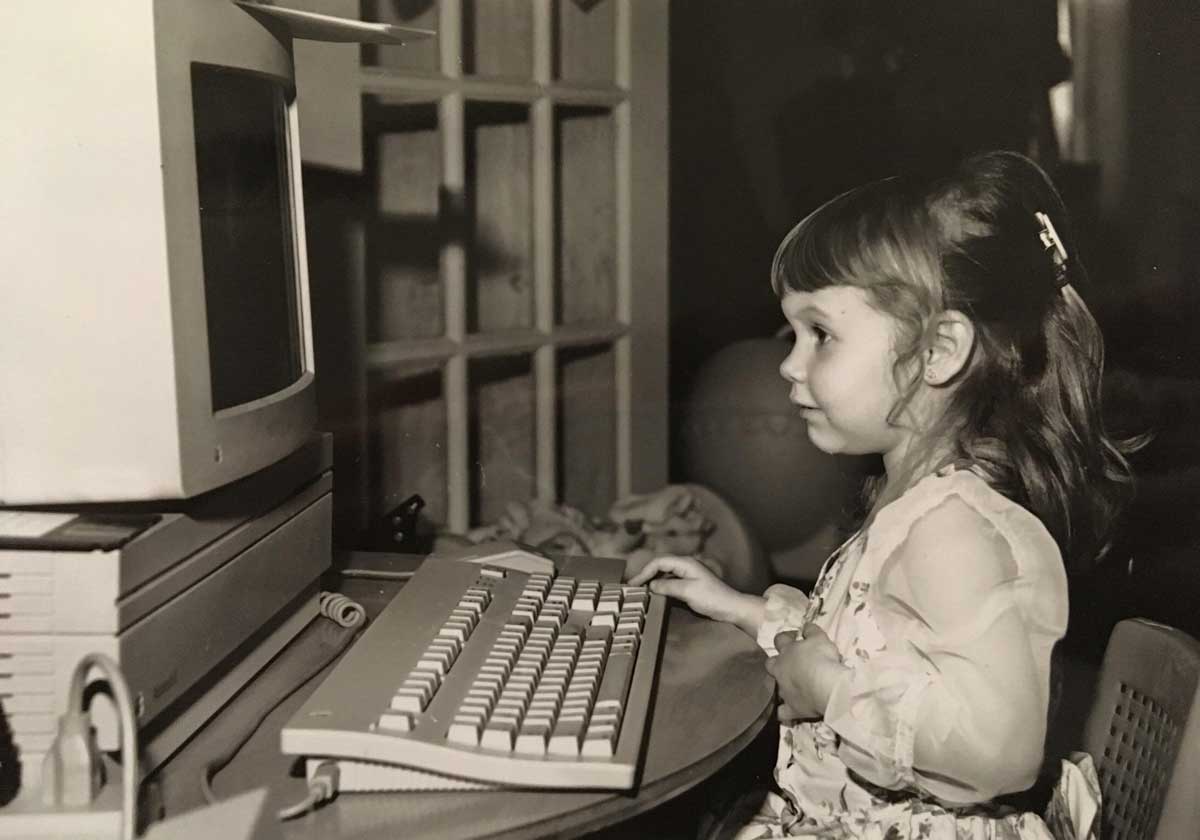 A young girl sits at a desk, using an old-fashioned computer with a large monitor and a keyboard. The photo is in black and white, giving it a nostalgic, vintage feel.