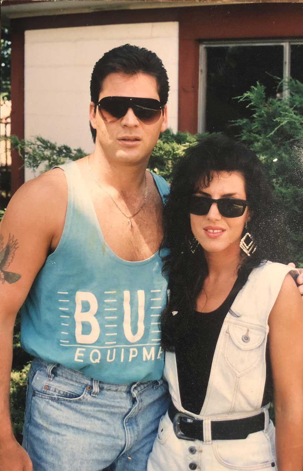 A man and a woman with dark hair and sunglasses pose together outside. The man wears a blue tank top and gold chain; the woman wears a white denim vest and large earrings. Both have a confident, 1980s-style appearance.