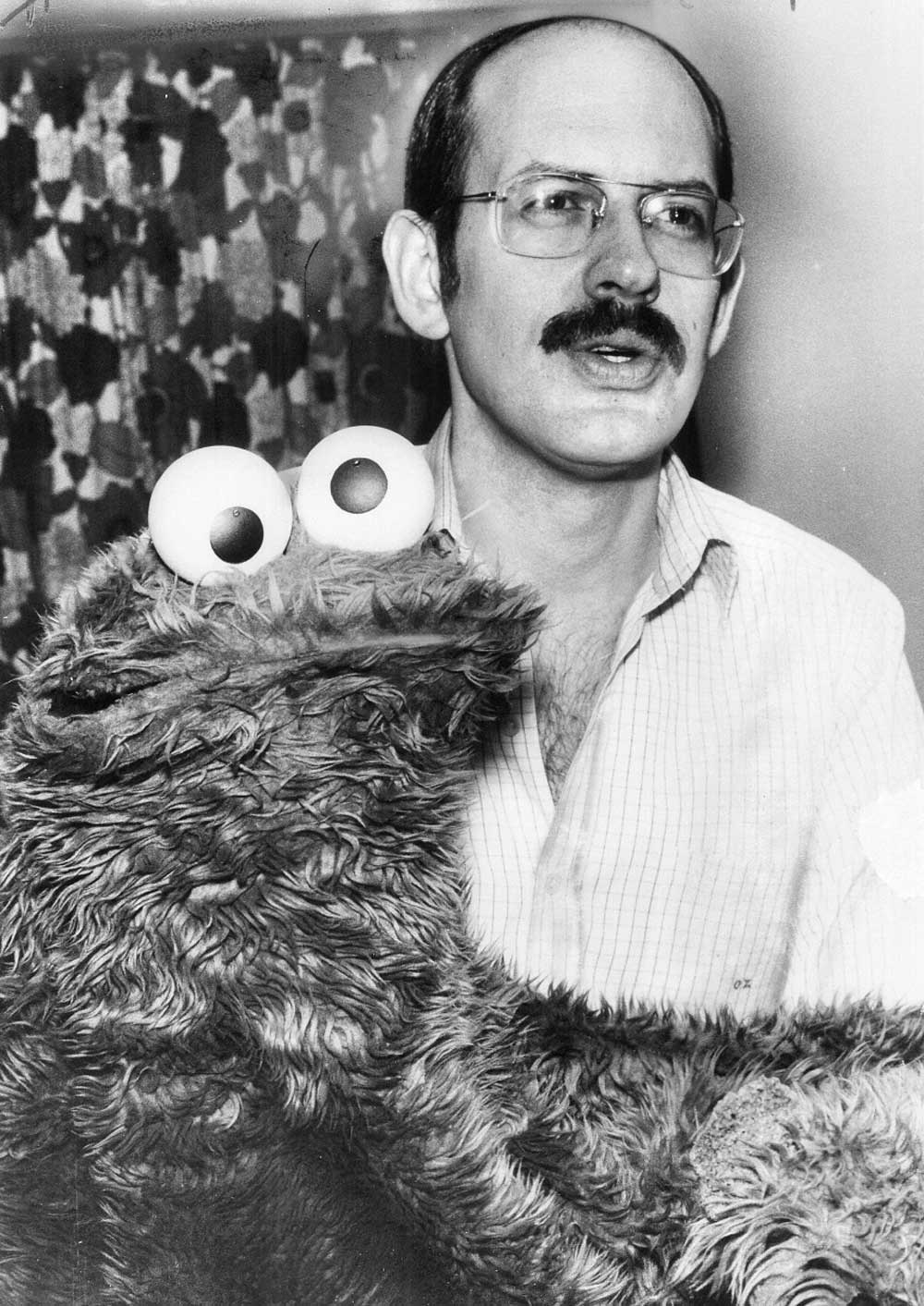 A man with glasses and a mustache poses with a large, fuzzy puppet resembling Cookie Monster. The man wears a light-colored shirt, and the background features a patterned curtain.