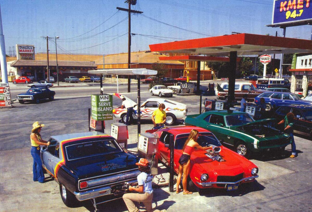 People in 1970s-style clothing pump gas into classic muscle cars at a busy retro gas station. Vintage buildings, parked cars, and vibrant colors add to the nostalgic atmosphere.