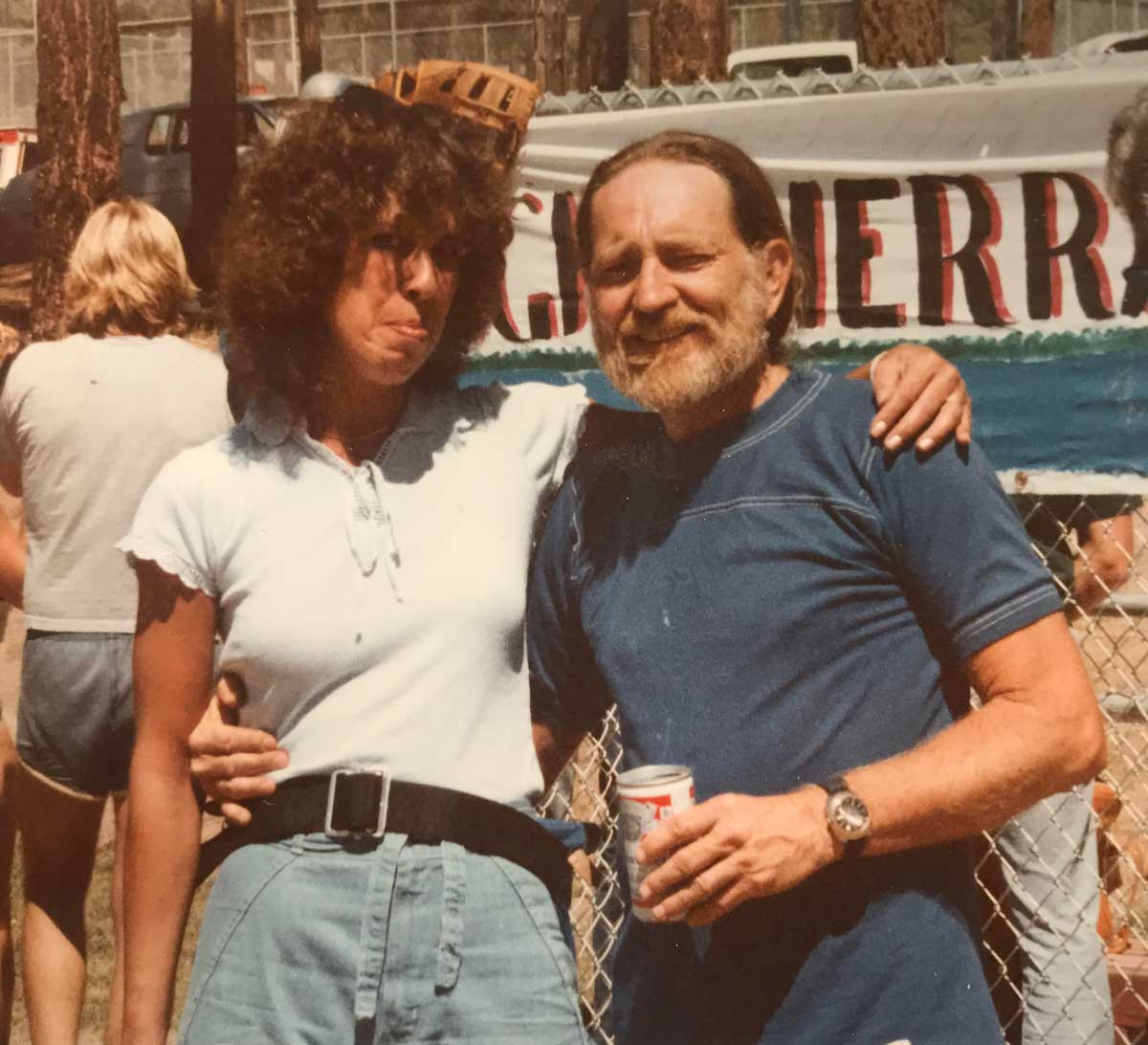 A woman and a man stand arm-in-arm, smiling outdoors by a chain-link fence. The woman wears a light shirt and denim shorts, the man holds a can and wears a blue shirt. A banner and people are visible in the background.
