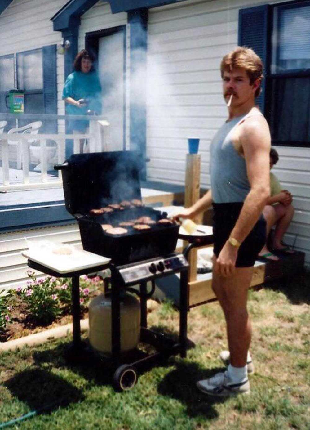 A man in a tank top and shorts grills burgers outside a house, holding a spatula with a cigarette in his mouth. Smoke rises from the grill. A woman stands on the porch and a child sits nearby.