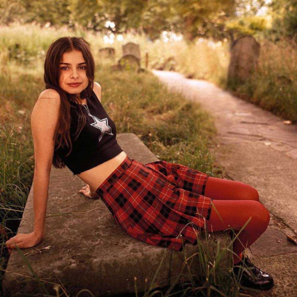 A young woman with long brown hair, wearing a sleeveless black top with a star, red plaid skirt, and red tights, sits on a stone slab outdoors along a path with grass and trees in the background.