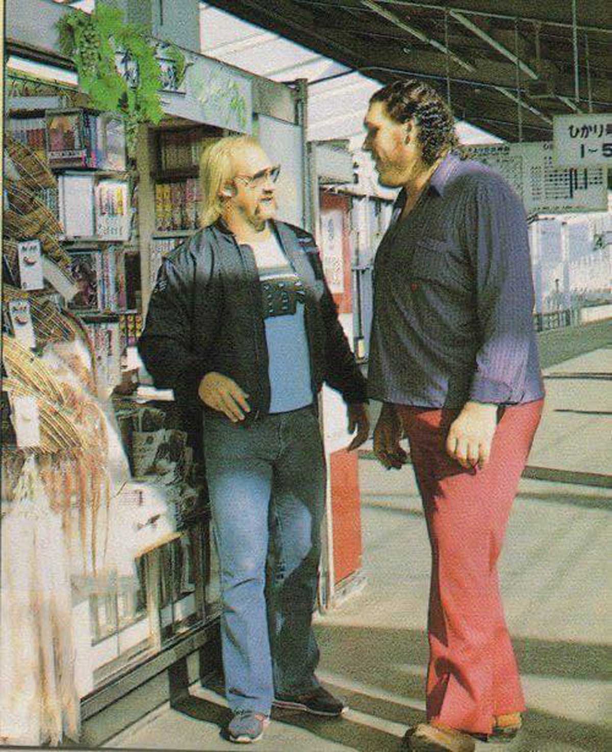 Hulk Hogan and Andre the Giant talk at an outdoor newsstand in Japan. Andre is much taller and wearing red pants and a striped shirt, while Hulk Hogan wears sunglasses, a black jacket, and blue jeans. Sunlight casts shadows on the ground.