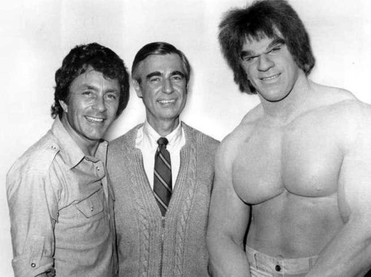 Three men pose together. The man on the left wears a button-up shirt, the man in the center wears a cardigan and tie, and the man on the right is shirtless, muscular, and has wild hair resembling a comic book character.