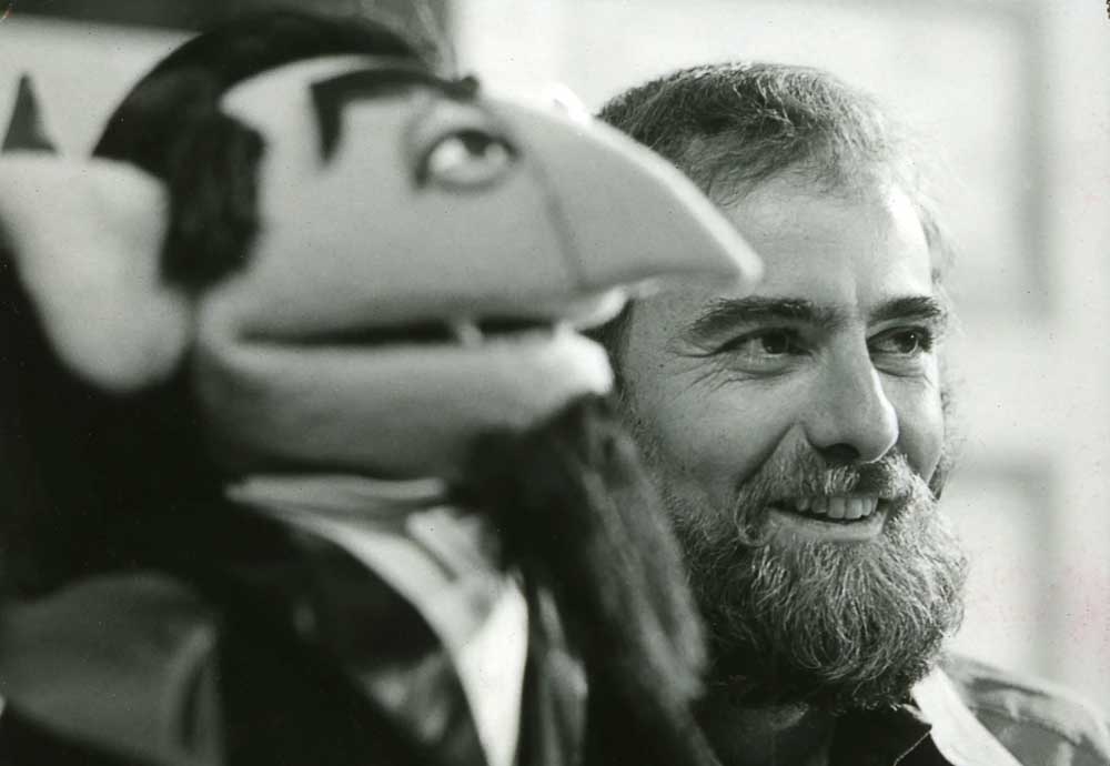 A black-and-white photo showing a smiling, bearded man next to a puppet with a long nose, pointed ears, and a goatee, both appearing to look in the same direction.