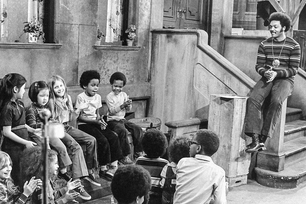 A group of children sit on steps and listen to an adult man speaking on an urban set resembling a stoop. The scene appears lively, with some children smiling and clapping. The setting looks like a TV show or classroom.