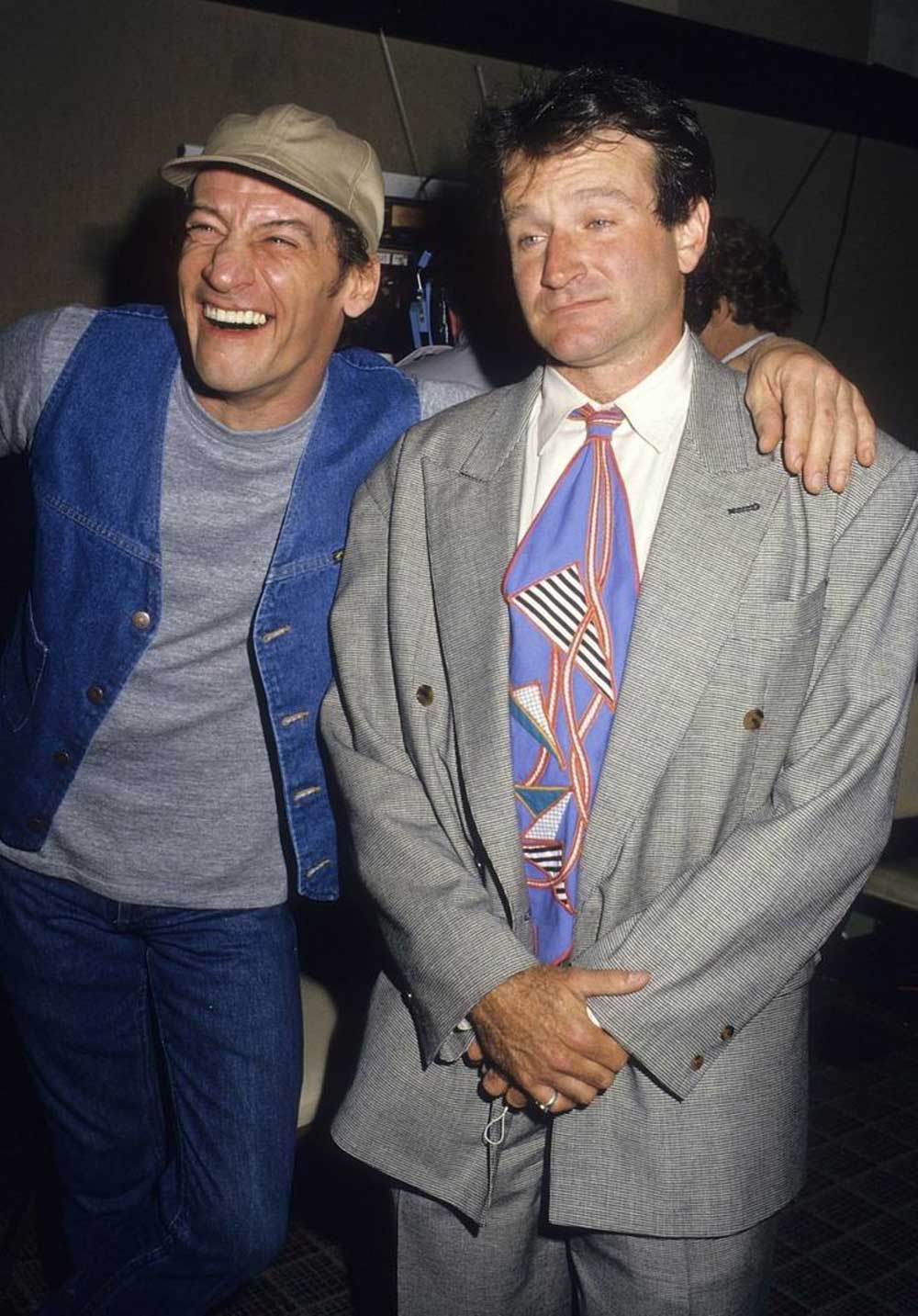 Jim Varney and Robin Williams stand side by side; one, in a cap and denim vest, laughs with his arm around the other, who wears a gray suit and colorful tie, and looks at the camera with a reserved expression.