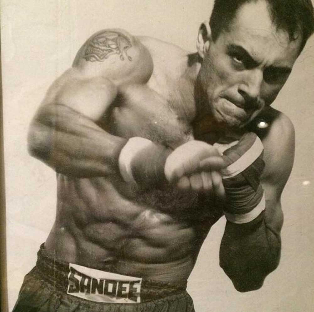 A muscular male boxer with wrapped fists throws a punch, wearing shorts labeled SANDERS. He has a tattoo on his upper arm and an intense expression on his face in this black-and-white photo.