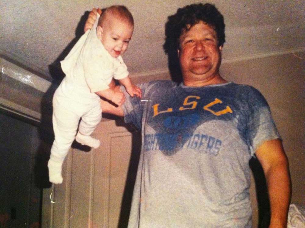 A smiling man in an LSU T-shirt holds a laughing baby up by the back of its shirt with one hand inside a room. The baby appears happy and excited, with arms and legs extended.