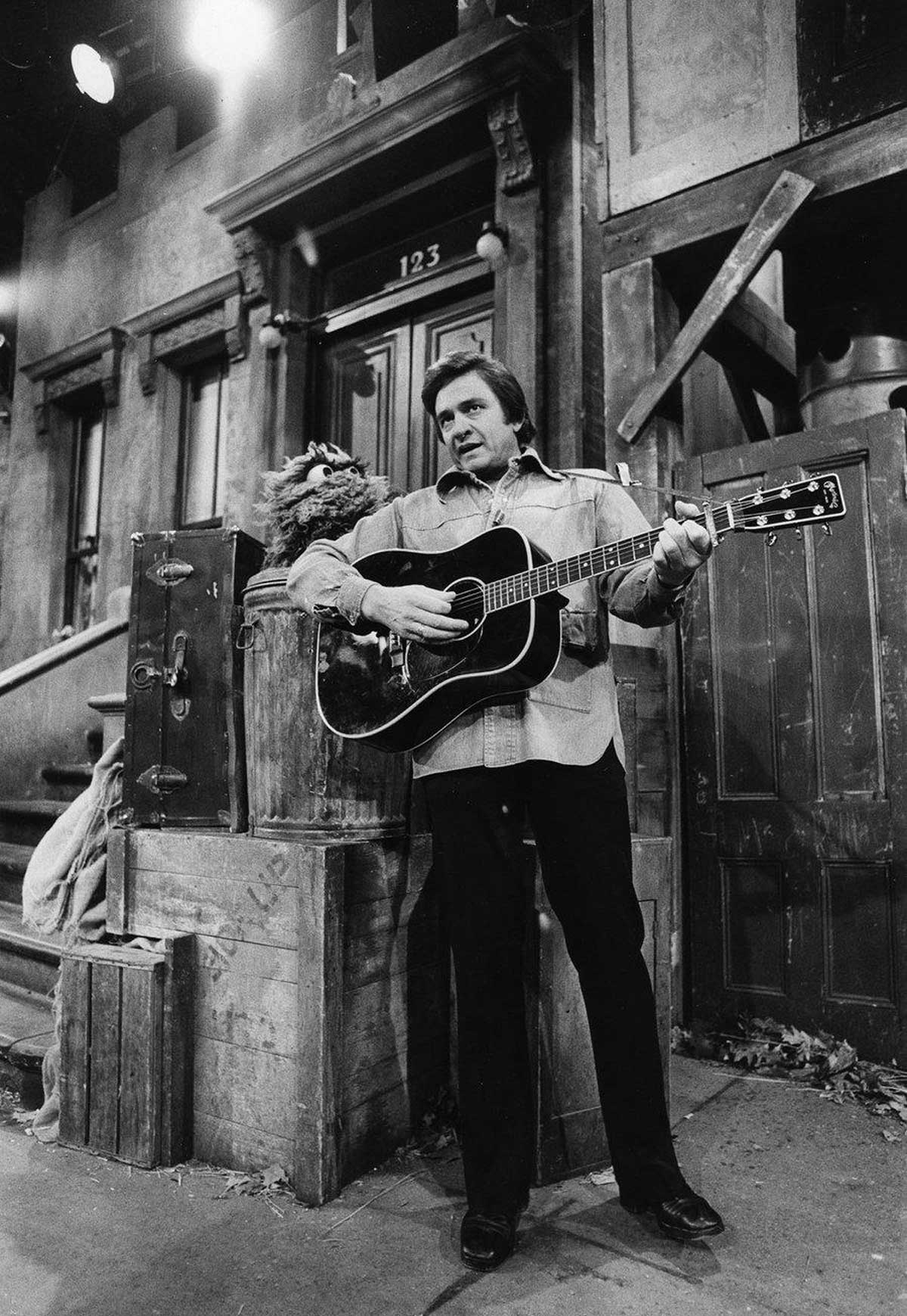A man stands playing an acoustic guitar on a city stoop set, with a puppet character peeking out of a trash can beside him. The scene is in black and white and appears to be from a television show.