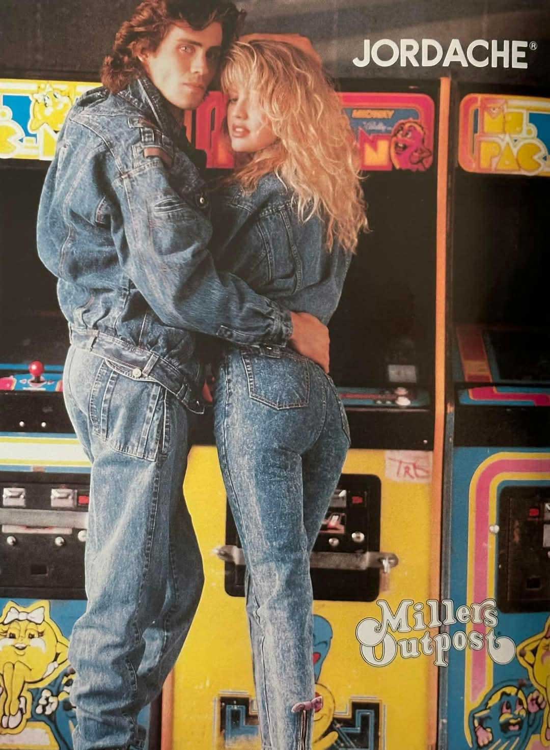 A man and woman wearing matching blue denim outfits pose in front of retro arcade game machines, including Ms. Pac-Man. The image features the Jordache logo in the top right corner and Miles Outpost in the bottom right.