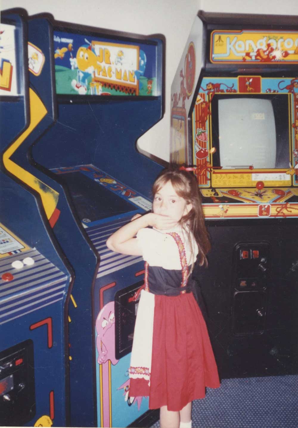 A young girl in a red and white dress stands in front of classic arcade machines, including Jr. Pac-Man and Kangaroo, looking over her shoulder at the camera.
