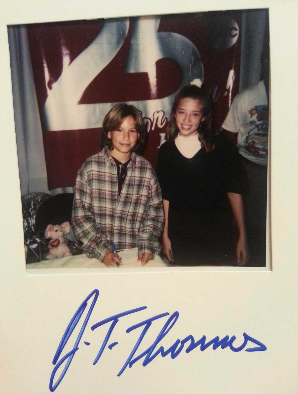 Two children, one in a plaid shirt and the other in a dark dress, smile at the camera. They stand in front of a banner with 25 on it. The photo has a large blue autograph at the bottom.