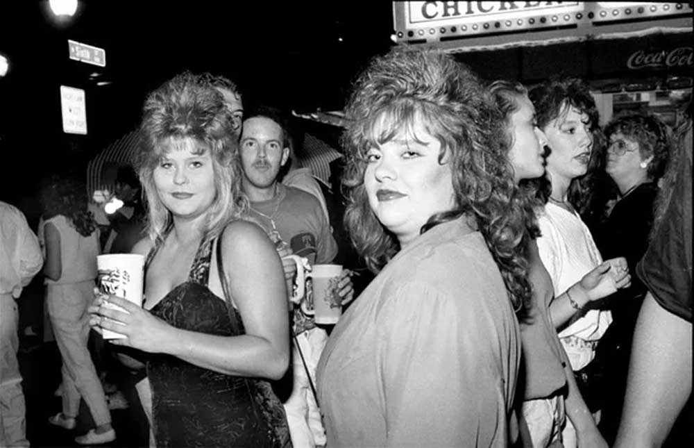 A group of people with big, teased hairstyles and 1980s fashion stand outside at night, holding drinks and looking at the camera. A restaurant sign and a Coca-Cola logo are visible in the background.