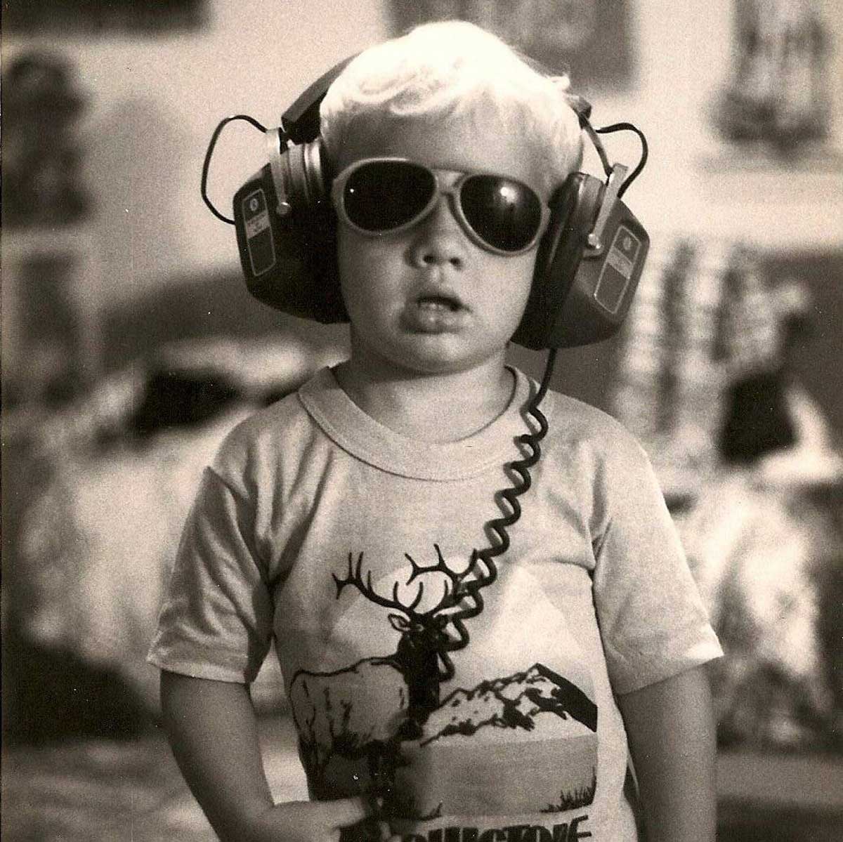 A young child wearing large headphones and sunglasses stands indoors, looking serious. The child has light hair and is wearing a t-shirt with a graphic of a deer and mountains. The photo is black and white.