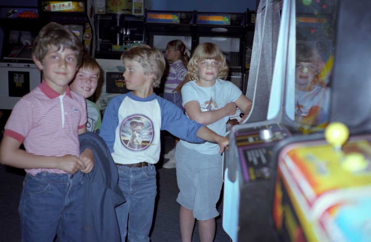 Four children stand in front of an arcade game, smiling and looking around. Two have their hands on the game controls, and arcade machines line the background. Their reflections are visible on the game screen.